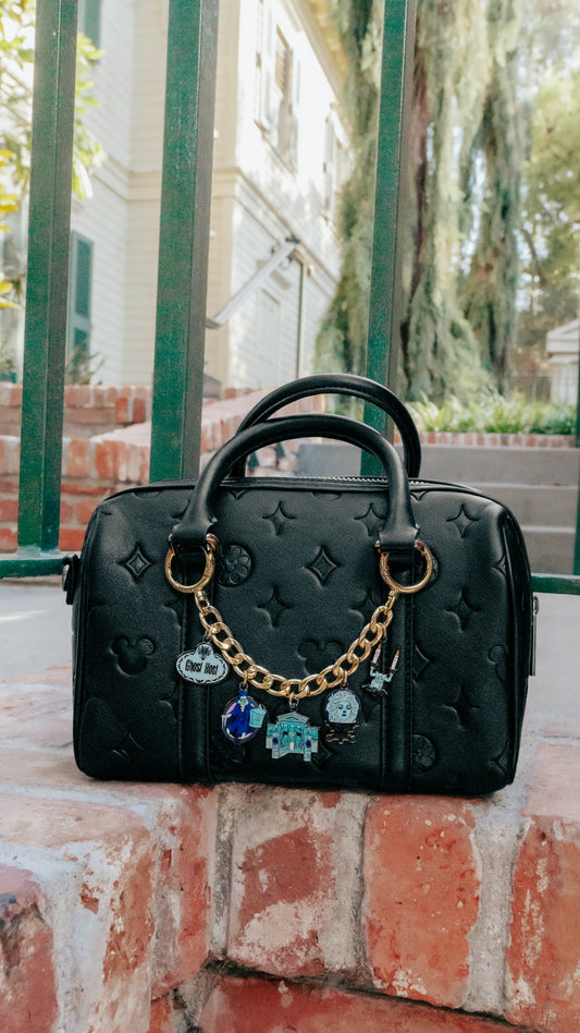 A black embossed leather handbag with gold-tone hardware and a decorative chain charm, resting on a brick ledge outdoors in front of a green metal fence.