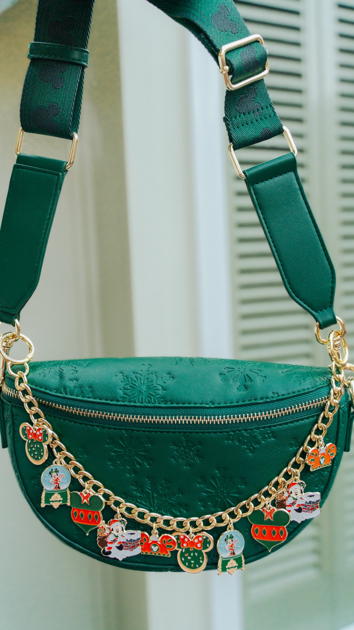 A green faux-leather fanny pack with a gold zipper and a decorative gold chain adorned with colorful Christmas-themed enamel charms, shown against a soft-focus background of white shutters.