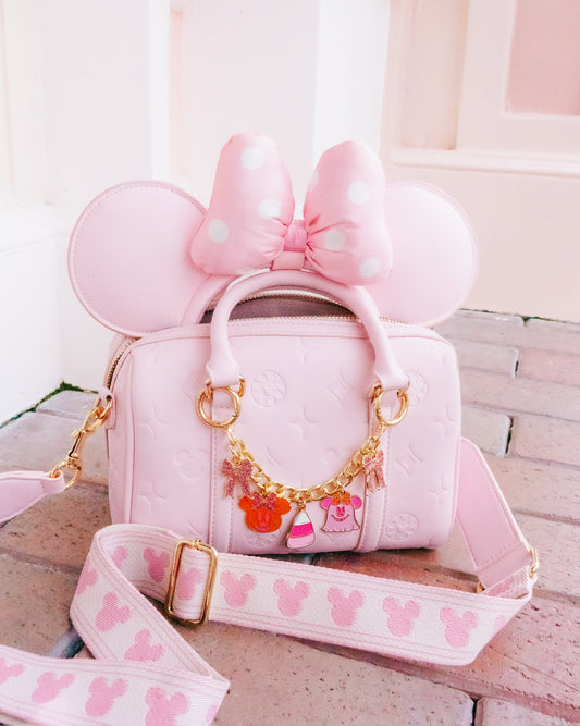 A pink Minnie Mouse-themed handbag with embossed floral details, a large bow on the top handle, mouse ears, and a decorative gold chain with character charms. A matching pink shoulder strap with a repeating Mickey Mouse head pattern is draped in front on a light stone surface.