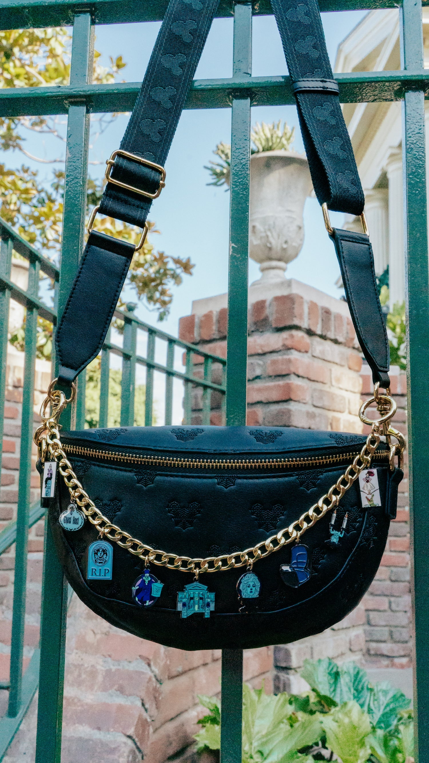 A black faux-leather crescent-shaped shoulder bag with embossed detailing, a gold zipper, a gold chain strap, and hanging blue and white charms, photographed outdoors against a green metal fence and brick wall.
