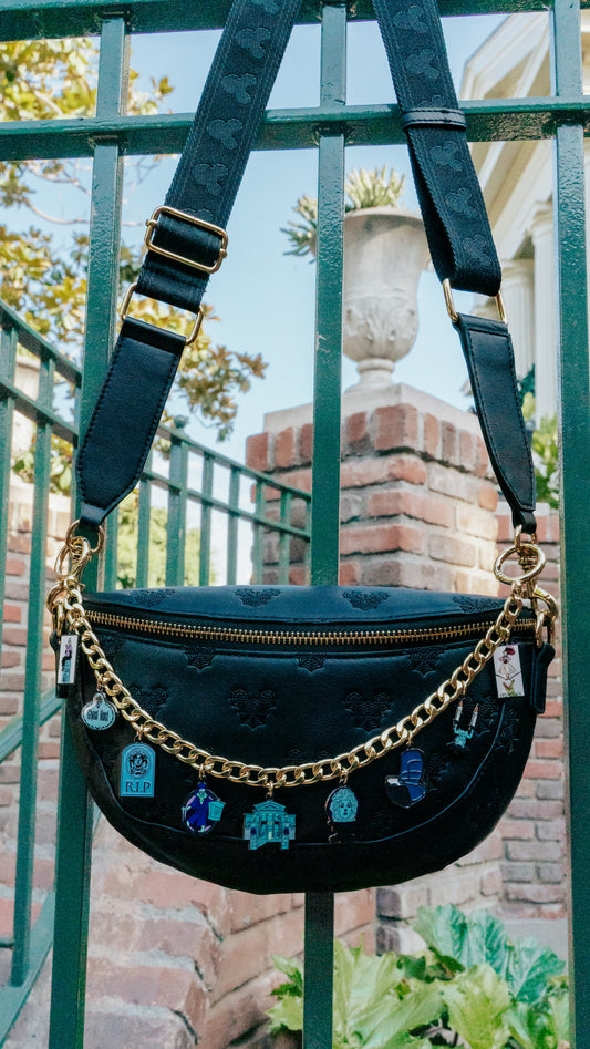 A black faux-leather crescent-shaped shoulder bag with embossed detailing, a gold zipper, a gold chain strap, and hanging blue and white charms, photographed outdoors against a green metal fence and brick wall.