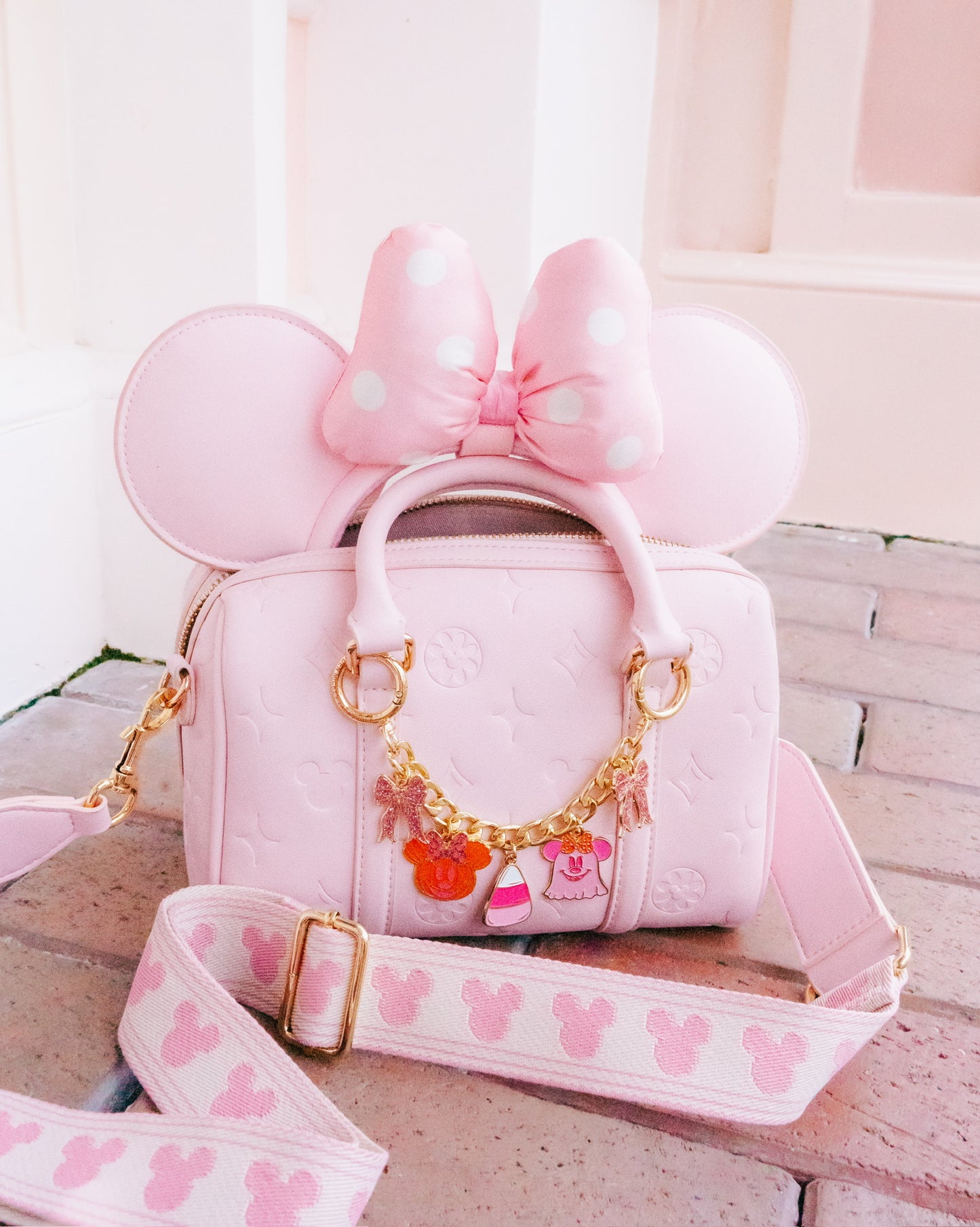 A pink Minnie Mouse-themed handbag with embossed floral details, a large bow on the top handle, mouse ears, and a decorative gold chain with character charms. A matching pink shoulder strap with a repeating Mickey Mouse head pattern is draped in front on a light stone surface.