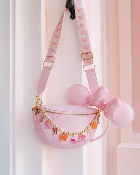A pastel pink crescent-shaped shoulder bag with gold hardware, featuring a large pink bow, round mouse-ear-like side decorations, and a patterned strap, hanging against a white door frame.