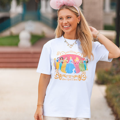 A woman wearing a white graphic t-shirt with a colorful Disney Princess print, white lace shorts, and a pink Minnie Mouse headband, standing outdoors on a paved path with a blurred resort background.