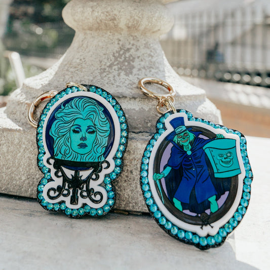 Two decorative keychains with colorful illustrated designs and turquoise rhinestone trim, displayed in front of a large stone urn outdoors.