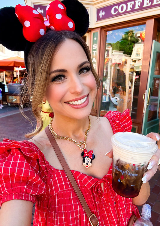 Woman in a red dress and Minnie Mouse ears holding a coffee cup in front of a Disney store.